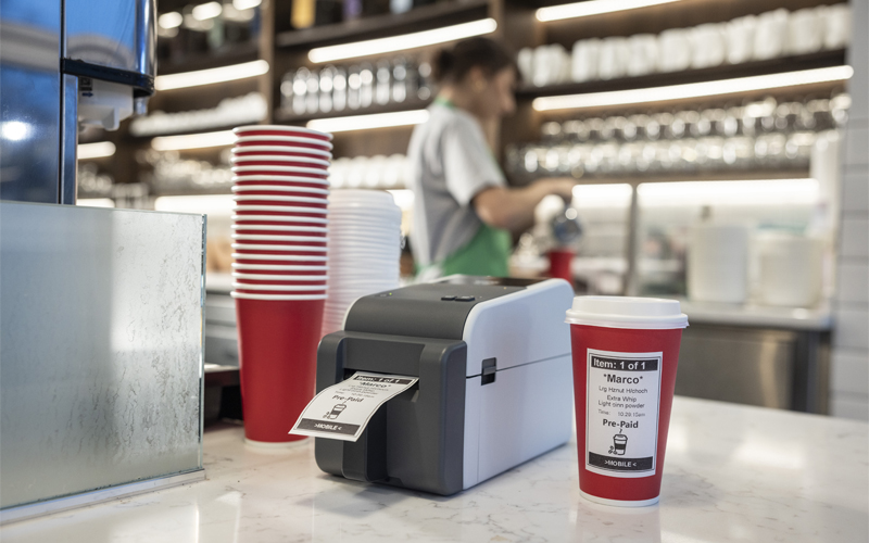 Caffetteria con donna sullo sfondo e una stampante per etichette e tazze da caffè in primo piano