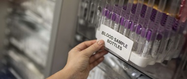 A selection of blood sample bottles have been labelled in a healthcare or hospital testing environment. The sticky label is being applied by a member of the medical staff. 